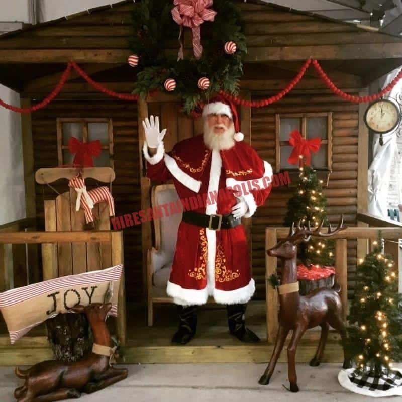 Real bearded Santa for hire in Indianapolis waving from a cabin doorway with wooden reindeer.