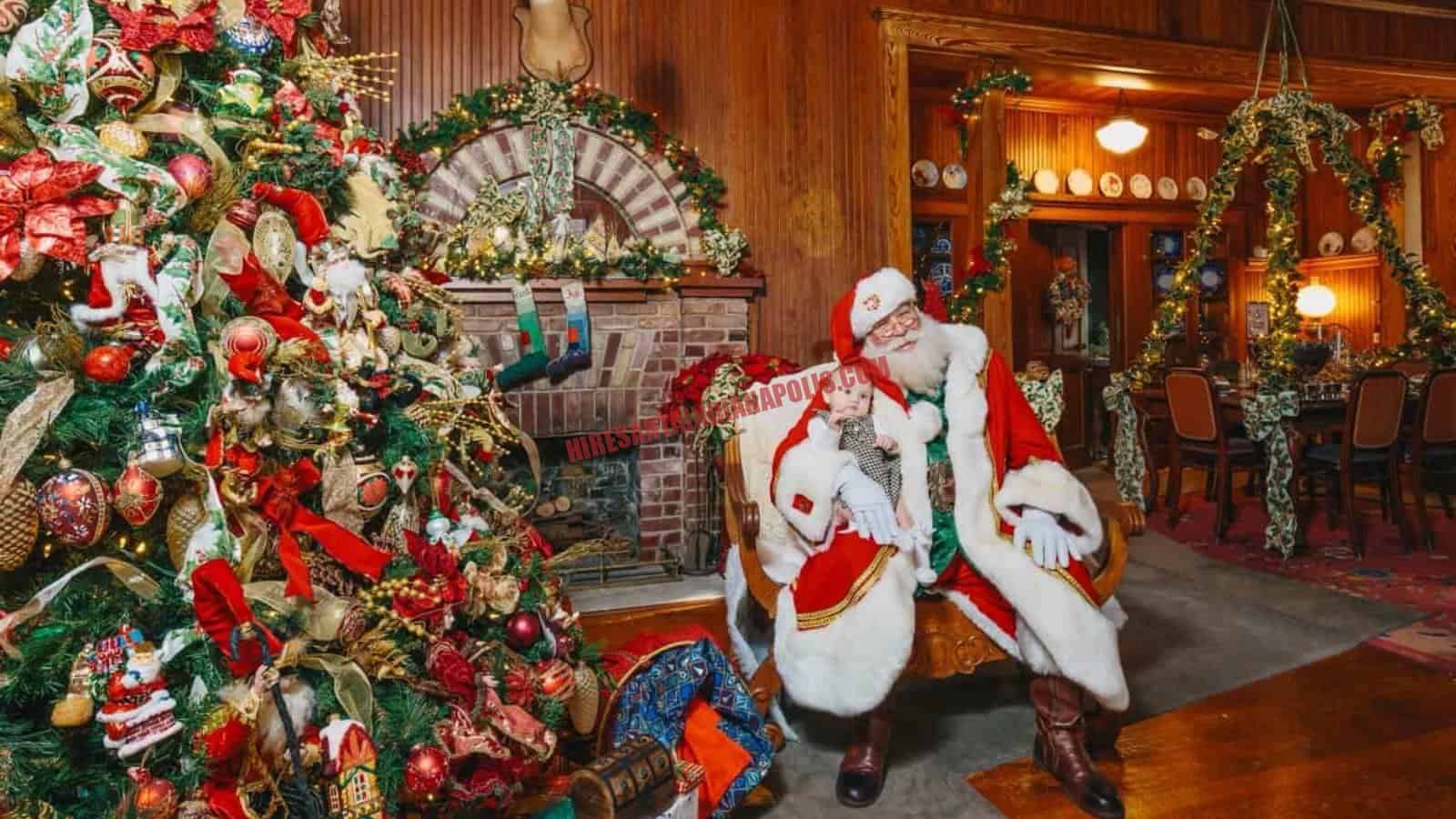 Santa Claus for hire in Indianapolis, IN seated by a fireplace and tree, holding a baby.