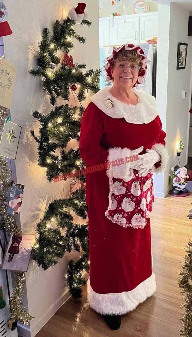 Mrs. Claus for hire in Indianapolis standing in a hallway by a slim lit tree in a red dress and printed apron.