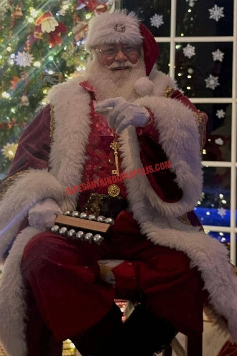 Santa for hire in Fishers, IN: Santa seated with sleigh bells by a tree and a night window.